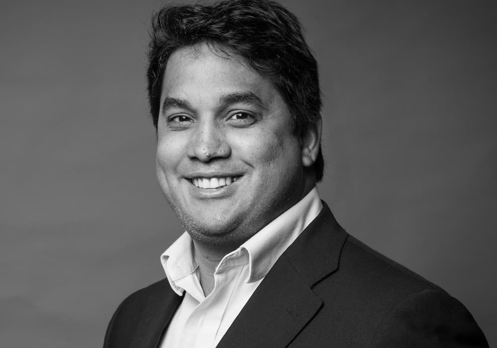 HugoAzevedo - crop SQSH Black and white photo of Hugo Azevedo's face and shoulders wearing a white button up and dark jacket, smiling with his teeth.